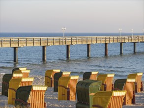 Colourful beach chairs stand on the sandy beach in front of a long wooden jetty over the calm sea,