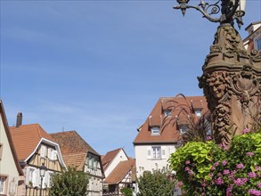 Image of a historic street lamp with half-timbered houses and flowers in the background,