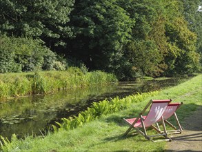 Two red deckchairs stand on the banks of a quiet river, surrounded by lush forest and green plants,