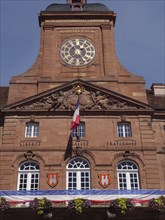 Close-up of a historic clock tower with French flag and richly decorated facade under a blue sky,