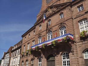 An imposing historic building, decorated with flowers and French flags, under a bright blue sky,