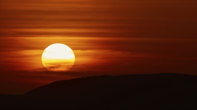 The setting sun breaks through dramatic layers of cloud and illuminates the silhouette of a hill,