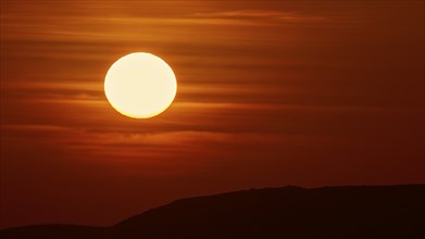 A large, bright ball of sunlight in the sky sinks behind the dark silhouettes of hills, offshore