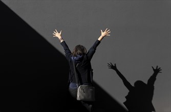 Woman Standing with Arms Raised Against a Grey Wall with Diagonal Shadow in a Sunny Day in