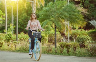 Beautiful and smiling young woman riding a bicycle in a nice park, looking at camera. Cheerful