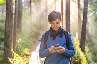 A boy stands on a forest path in the sunlight, carrying a rucksack and using his smartphone, AI