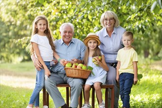 Grandparents and grandchildren outside in summer, all smiling, with a basket of apples. Harmonious