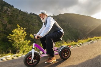 An elderly man rides a tricycle on a road in the middle of a natural environment and laughs