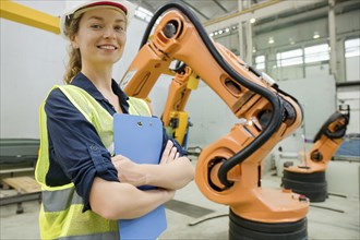 Woman with helmet and yellow safety waistcoat in a factory next to an orange robot arm, AI and