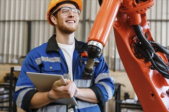 A worker in uniform and hard hat smiles while holding a tablet and standing next to a large robotic