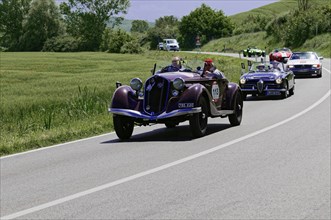 Purple vintage racing car driving on a country road, followed by other classic cars, vintage car,