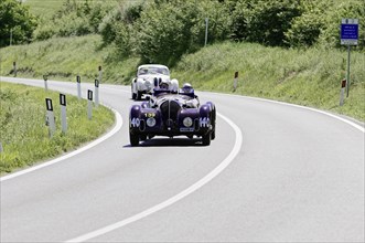 Purple vintage racing car and other vintage cars on a country road, surrounded by green nature,