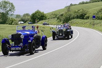 Blue vintage racing car on a winding country road, followed by other vintage racing cars, vintage