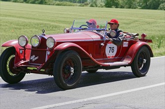 Two people driving in a red vintage racing car on a country road, surrounded by green nature,