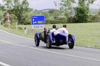 Blue vintage car with two people driving along a country road. Road sign to Siena visible, classic