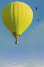 A bright yellow hot air balloon floating in a clear blue sky with smaller colorful balloons in the