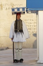 Evzone in traditional dress stands next to a guard hut under a sunshade, Parliament building, Tomb