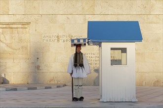 Evzone in traditional dress stands next to a guard hut under a sunshade, Parliament building, Tomb