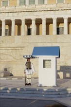 Evzone in traditional dress stands next to a guard hut under a sunshade, Parliament building, Tomb