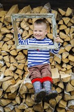 A smiling boy in a striped shirt and boots sits on a woodpile, holding a rustic, weathered frame