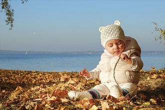 A baby in a white beanie and pink jacket sits among fallen autumn leaves by a lake, focusing on a