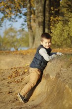 A young boy smiling and climbing a dirt slope in an autumn forest setting with colorful fall