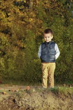 A young boy in a blue vest stands on a grassy area surrounded by autumn leaves and trees, Belarus