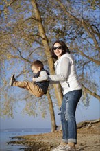 A woman lifting a young boy near a lake on a sunny autumn day with trees and blue sky in the