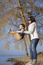 Woman lifting a smiling child near a lake with autumn trees in the background, Belarus.Minsk