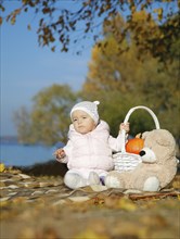 A baby sitting on a blanket outdoors in autumn with a teddy bear, a basket containing a pumpkin and