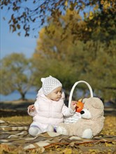 Baby in a pink jacket and white hat sitting on a blanket with a teddy bear and basket in an autumn