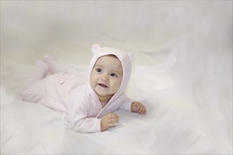 A baby in a pink outfit with a hood featuring bear ears lying on a white background, looking up and