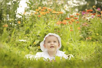 A smiling baby wearing a white hat sits amidst a garden filled with colorful flowers and lush