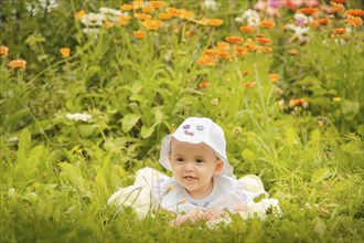 A baby wearing a hat lying in the grass surrounded by colorful flowers on a sunny day, Belarus