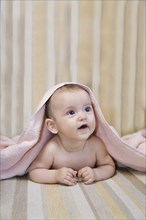 A baby lying on a striped surface, partially covered with a pink towel, looking up with an adorable