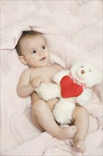 A baby lying on a pink blanket holding a white teddy bear with a red heart