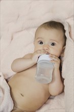 An infant lies on a pink blanket, holding a bottle with milk, looking curiously ahead