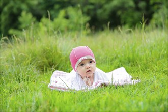 Baby lying on a white blanket in the grass, wearing a pink hat, surrounded by green foliage,