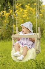 A baby sitting on a swing in a lush garden with yellow flowers, wearing a hat and dress while