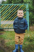 Young boy wearing a blue jacket and boots stands outdoors near a green gate.Belarus.Minsk