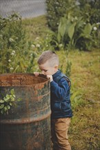 A young boy in a blue jacket and brown pants peers into a rusted metal barrel in a grassy outdoor