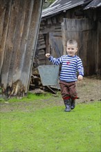 A boy carrying a metal bucket, wearing a striped shirt, brown pants, and rubber boots, walking on