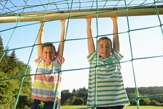 Two boys hanging from a goal post net, smiling, against a backdrop of greenery and a bright sunny