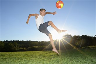 A boy jumping in the air about to kick a soccer ball on a grassy field during a sunny day, Bavaria