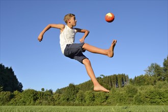 A boy mid-air kicking a soccer ball in a grassy field under a clear blue sky, Bavaria