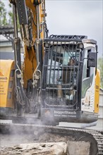 Excavator during demolition, breaking up concrete. The driver is visible in the cab, demolition