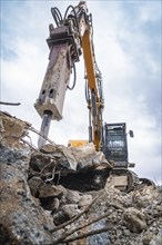 View of an excavator from below, breaking up concrete on a construction site. The sky is cloudy,