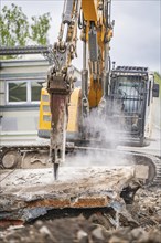 Excavator breaking concrete on a construction site, while a building can be seen in the background.