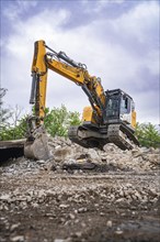 An excavator lifts large pieces of rubble on a construction site, with cloudy sky in the