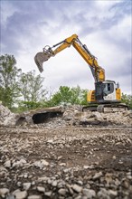 An excavator on a building site lifts rubble. Trees and sky in the background, demolition site,